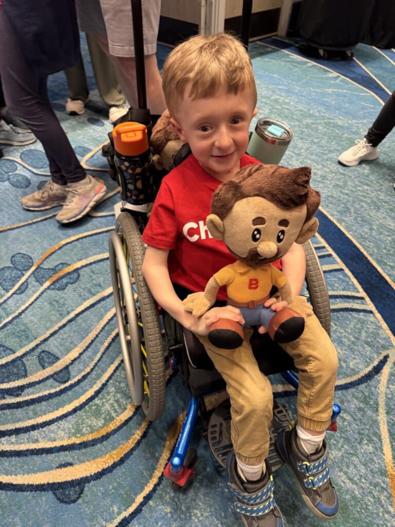 Young child in a wheelchair wearing a red shirt with white text, tan pants, and blue sneakers holding a plush doll with brown hair, beard, and a yellow shirt with a red "B" on it.