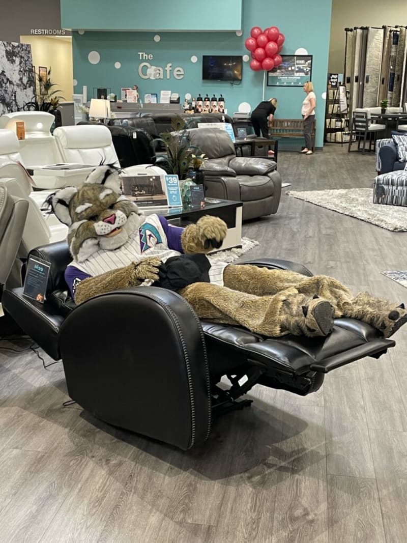 A person in a bobcat mascot costume wearing a baseball jersey reclines on a black leather power recliner chair in a furniture store showroom with sofas and a cafe area in the background.
