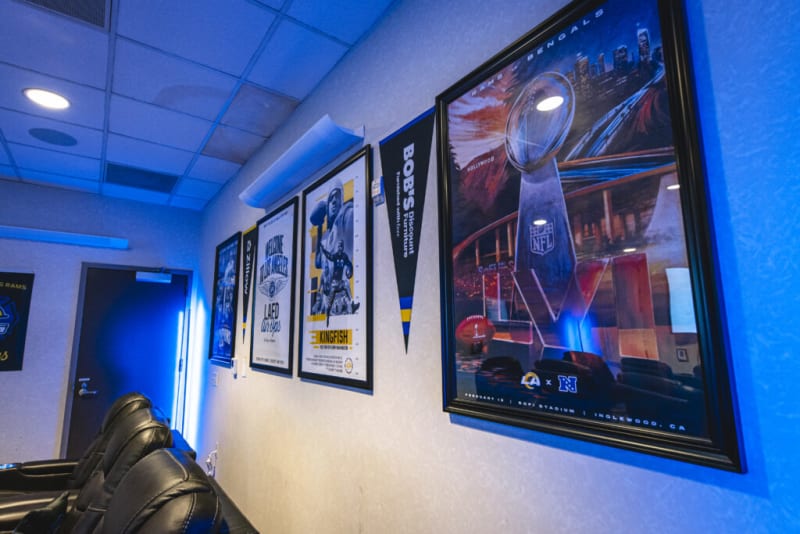 Framed sports memorabilia and posters on a wall with NFL and LA Rams themes, including a Super Bowl LVI poster and a "Welcome to the Valley" LAED Chiefs sign.