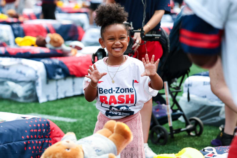 Child wearing a white "Bed-Zone BASH" t-shirt and pink skirt, standing near colorful bedding sets and plush toys on green turf.