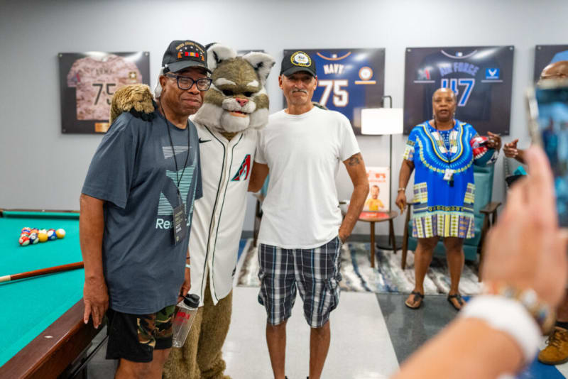 Three men stand next to a person in a furry Arizona Diamondbacks mascot costume near a pool table, with sports jerseys displayed on the wall.