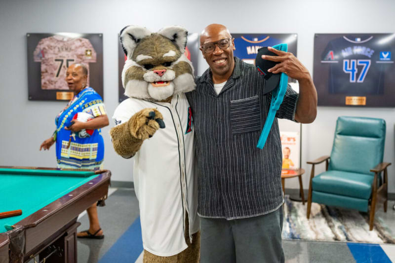 Man posing with a sports team mascot in a white jersey next to a pool table, with sports memorabilia and framed jerseys on the wall.