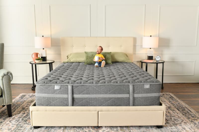 King-size bed with beige upholstered headboard and base, gray extra firm mattress, green pillows, two black round side tables with lamps, clock, and decor.