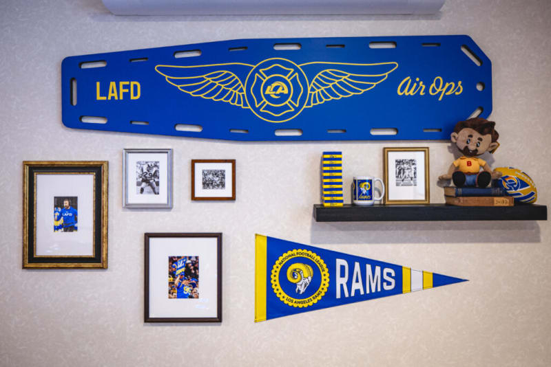 Blue and yellow LAFD Air Ops sign with wings logo, framed sports photos, Los Angeles Rams pennant, mug, and memorabilia on wall shelf.