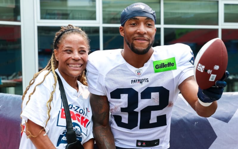 Two people stand together, one wearing a white New England Patriots football jersey with number 32 and a Gillette patch, holding a football. The other person wears a white T-shirt with blue and red text reading "No-Zoll Bash." They are outdoors near a building with large windows.