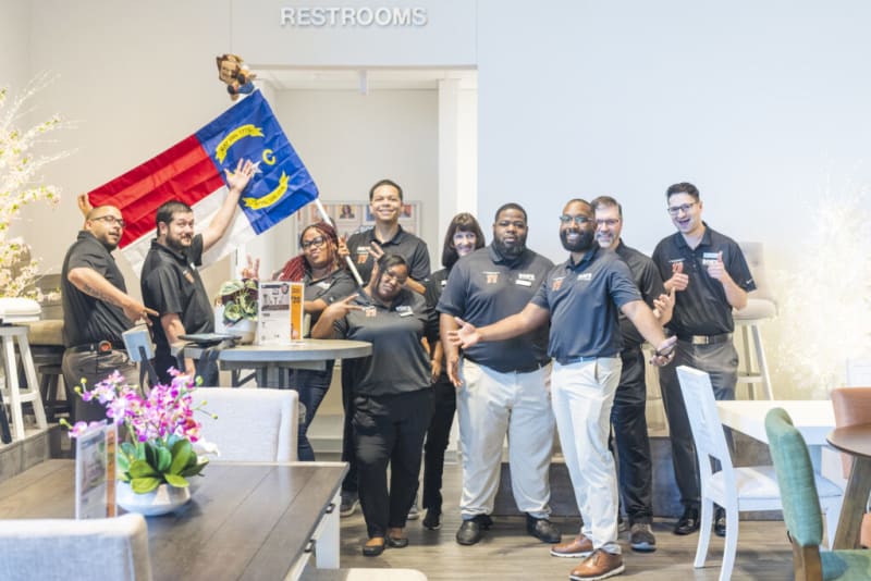 Group of people wearing black polo shirts and light-colored pants standing indoors near a table, one holding a North Carolina state flag, with a "RESTROOMS" sign above.