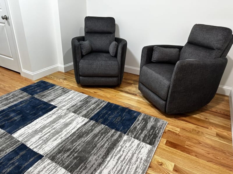 Two dark gray fabric upholstered recliner chairs with cushioned armrests and headrests on a hardwood floor next to a modern blue, gray, and white patterned area rug in a minimalist room.