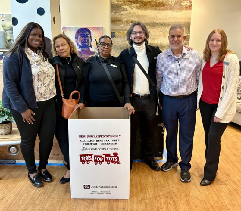 Group of six people standing indoors around a large white Toys for Tots donation box with text: "NEW, UNWRAPPED TOYS ONLY CAMPAIGN RUNS 1 OCTOBER THROUGH DECEMBER MARINE CORPS RESERVE TOYS FOR TOTS Welch Packaging Group MAKING A DIFFERENCE." The setting appears to be a community or charity event promoting toy donations.