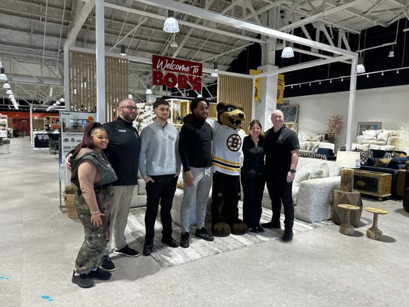 Group of six people standing inside a furniture store near a "Welcome to Bob's" sign, posing with a Boston Bruins mascot in a white and black jersey.