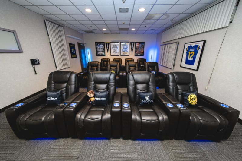 A modern home theater room with two rows of black leather recliner chairs featuring branded pillows from Zillow, eHomes, and LA Galaxy, framed sports memorabilia on the walls, and blue ambient lighting.