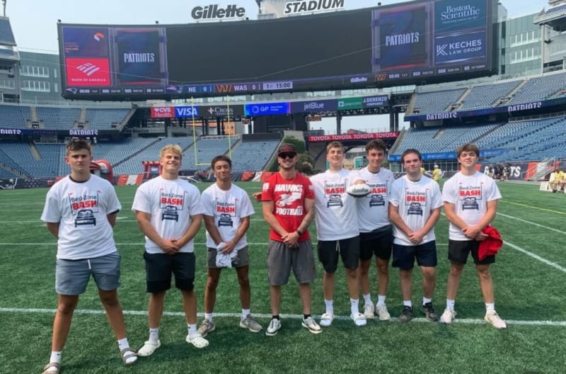 Group of eight men standing on Gillette Stadium field wearing Bed Zone Bash event t-shirts, shorts, and athletic shoes, with stadium seating and scoreboard in the background.