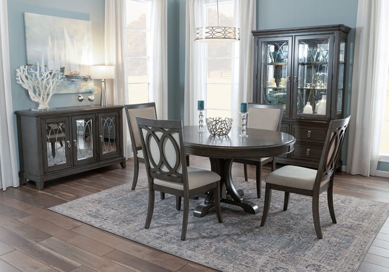 Dark wood dining room set with oval table, four cushioned chairs, matching sideboard and glass-front cabinet on patterned rug.