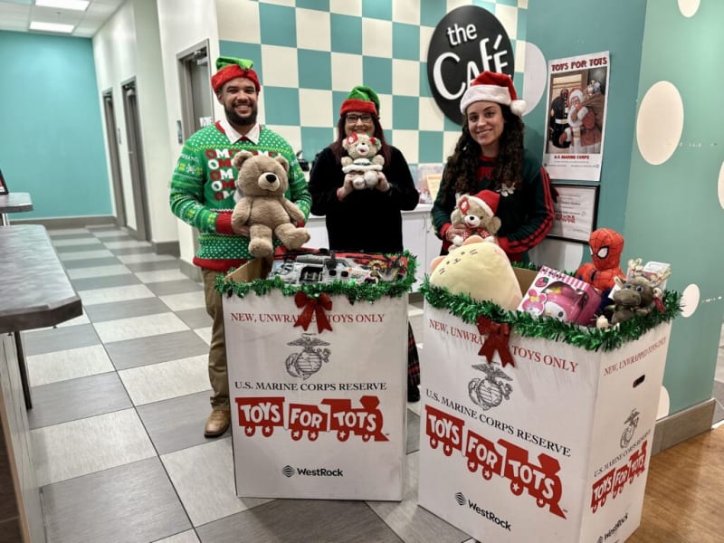 Three people in festive holiday attire stand behind two large white collection boxes labeled "TOYS FOR TOTS" with U.S. Marine Corps Reserve logos, filled with various stuffed animals and toys. The boxes also feature the text "NEW, UNWRAPPED TOYS ONLY" and "WestRock." The setting is a café with teal walls and checkered tiles.
