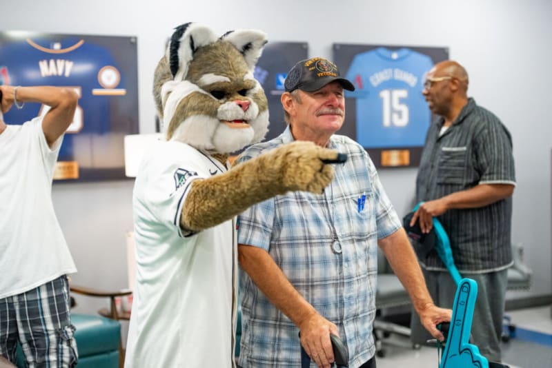 A person in a cat mascot costume wearing a white sports jersey points forward while standing next to a man in a plaid shirt and baseball cap holding a blue foam finger in a sports-themed room with framed jerseys on the wall.