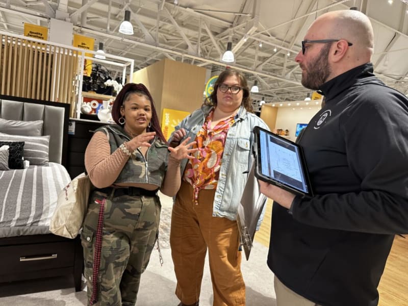 Three people in a furniture store discussing with a tablet; one person wears camouflage pants and a cropped vest, another wears brown pants and a denim jacket, and the third wears a black long-sleeve shirt holding a tablet displaying a floor plan.