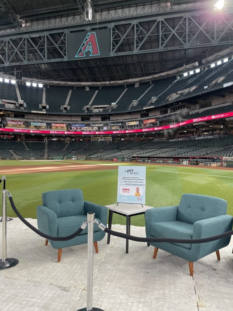 Two teal upholstered armchairs with wooden legs and a small white square table between them are set on a white platform inside a large empty baseball stadium. A sign on the table reads: "a gift for you. Supporter of Arizona Diamondbacks Foundation from Bob’s Discount Furniture. Amount $2,500 Shopping Spree. BOB'S DISCOUNT FURNITURE." The setting highlights a promotional event for Bob’s Discount Furniture at the Arizona Diamondbacks stadium.