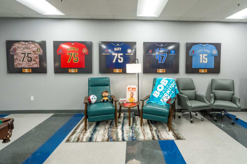 Wall display of five framed sports jerseys representing U.S. military branches: Army (75), Marines (75), Navy (75), Air Force (47), and Coast Guard (15), above a seating area with teal and gray chairs, a small table, and a floor lamp in a modern office space.