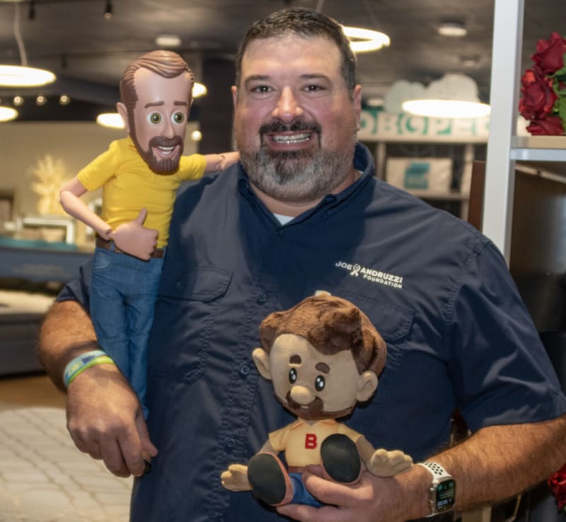 Man wearing a navy blue Joe Andruzzi Foundation shirt holding two plush dolls, one with a yellow shirt and blue pants and the other with a beige shirt with a red "B" and blue pants.