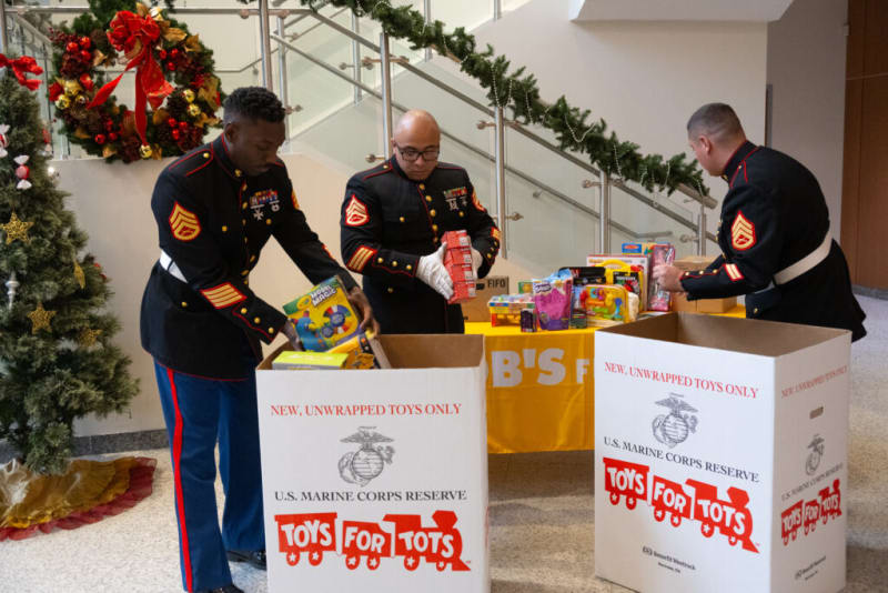 Three U.S. Marine Corps Reserve members in dress uniforms sorting new, unwrapped toys into large white "Toys for Tots" donation boxes next to a decorated Christmas tree and festive wreath.