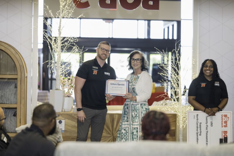 Three people stand inside a Bob's Discount Furniture store during a certificate presentation. The person on the left wears a black Bob's Discount Furniture polo shirt with a number 11 patch and gray pants, holding a "Certificate of Appreciation." The person in the middle wears a white and green patterned dress with a white cardigan. The person on the right wears a black Bob's Discount Furniture polo shirt and holds a large check that reads: "Bob's Outreach, $25,000, Supporting North Carolina Operators." Decorative white lighted trees and furniture are visible in the background.