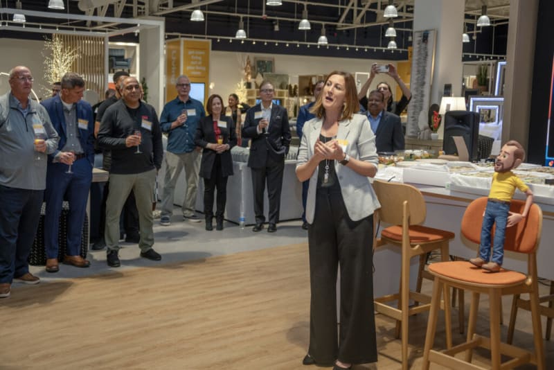 A woman in a light gray blazer and black pants stands in front of a group of people holding drinks at a social or networking event in a modern indoor venue with wood flooring and contemporary lighting.
