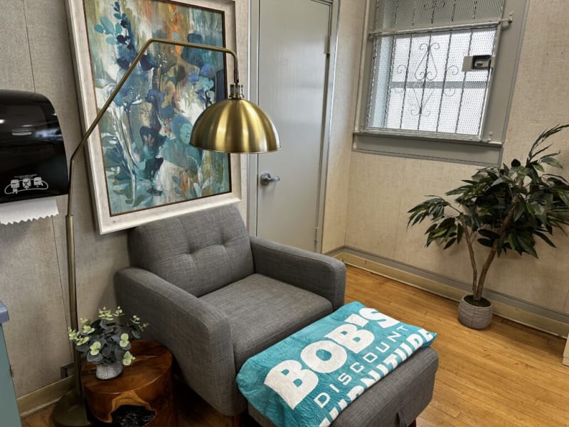 Gray upholstered armchair with matching ottoman, topped with a turquoise blanket displaying "BOB'S DISCOUNT FURNITURE," next to a brass floor lamp and decorative plants in a cozy room.
