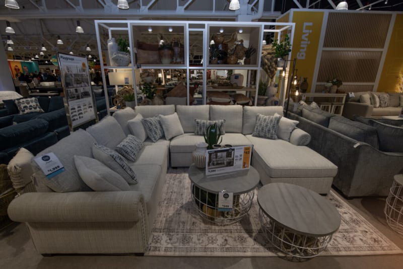 Large beige fabric sectional sofa with patterned cushions, two round wooden coffee tables with metal bases, displayed in a modern living room showroom. Price tags show $1,999 and $249. The background includes shelves with decorative items and a yellow wall labeled "Living."