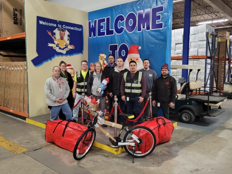 Group of warehouse employees standing in front of a blue banner with "WELCOME TO" text and a Santa Claus decoration, inside a storage area with boxed furniture and a golf cart. A red and black bicycle with large red bags is in the foreground.