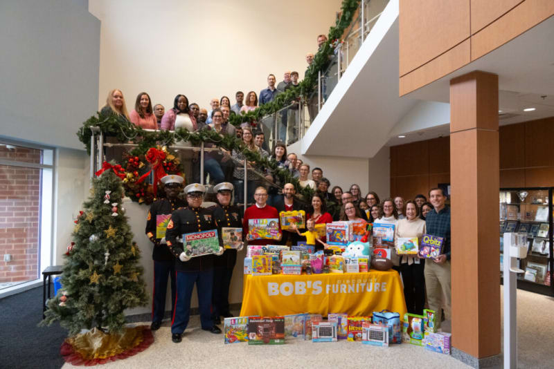 Group of people posing on a staircase and around a table with a yellow "Bob's Discount Furniture" tablecloth displaying various board games and toys, including Monopoly, Sorry, Life, and Q-bitz, next to a decorated Christmas tree with gold and red ornaments.