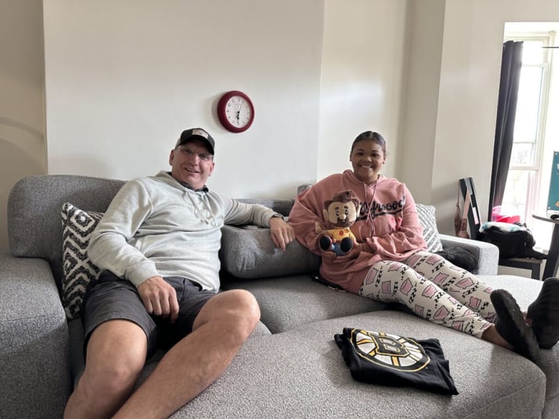 Two people sitting on a gray fabric sectional sofa with patterned pillows, one holding a plush monkey toy, a folded black Boston Bruins shirt on the ottoman, and a red wall clock showing 12:05.