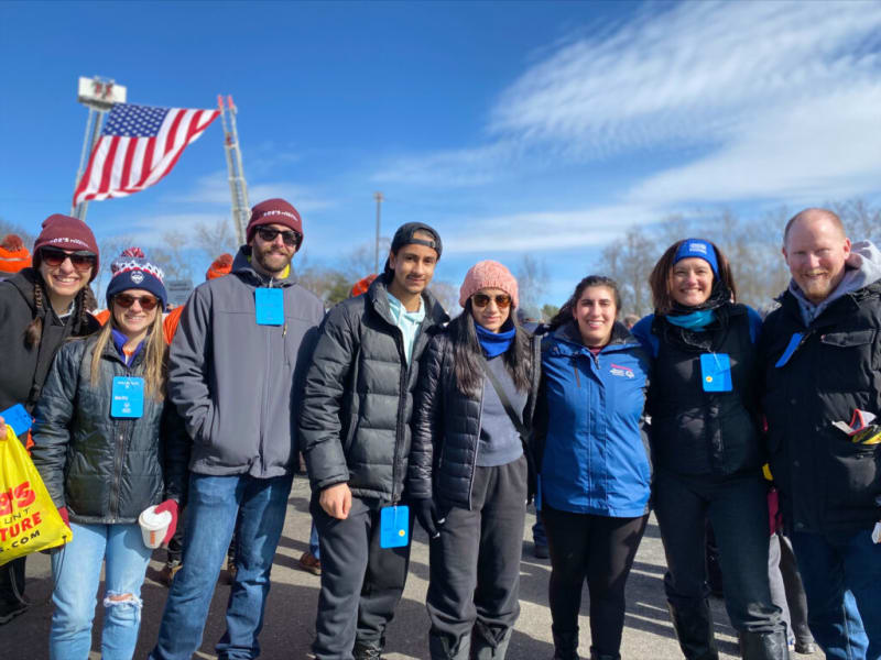 Group of people dressed in winter jackets and hats standing outdoors under a clear blue sky with a large American flag in the background, suggesting a community or public event.