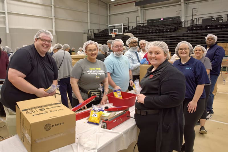 Volunteers wearing hairnets and casual clothing pack food items like rice and pasta into cardboard boxes on a table in a gymnasium, supporting a community food drive.