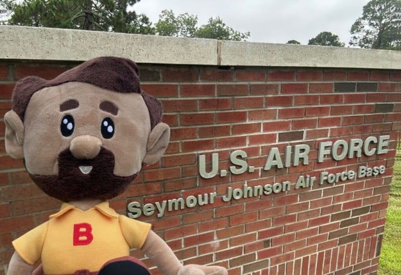 Plush doll with brown hair and beard wearing a yellow shirt with red "B" in front of a brick wall sign reading "U.S. AIR FORCE Seymour Johnson Air Force Base."