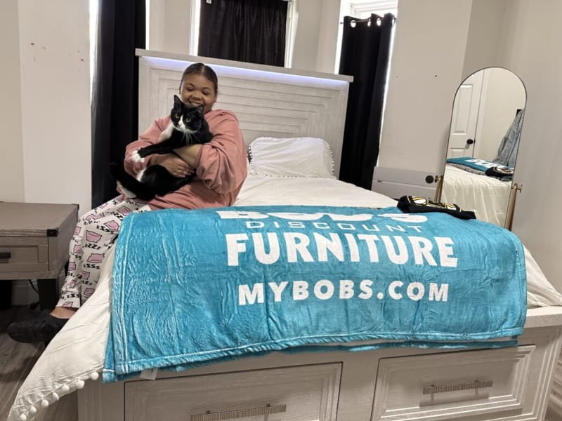 Person sitting on a white bed with a blue plush blanket draped over the footboard displaying "DISCOUNT FURNITURE MYBOBS.COM" in white text.