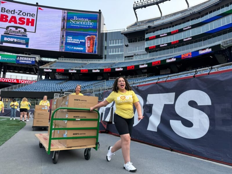 Volunteer in yellow T-shirt pushing a green-handled cart loaded with stacked cardboard boxes labeled "BOB'S FURNITURE" inside a large stadium with digital advertising screens displaying "Bed-Zone BASH," "Boston Scientific," and "BOB'S Discount Furniture."
