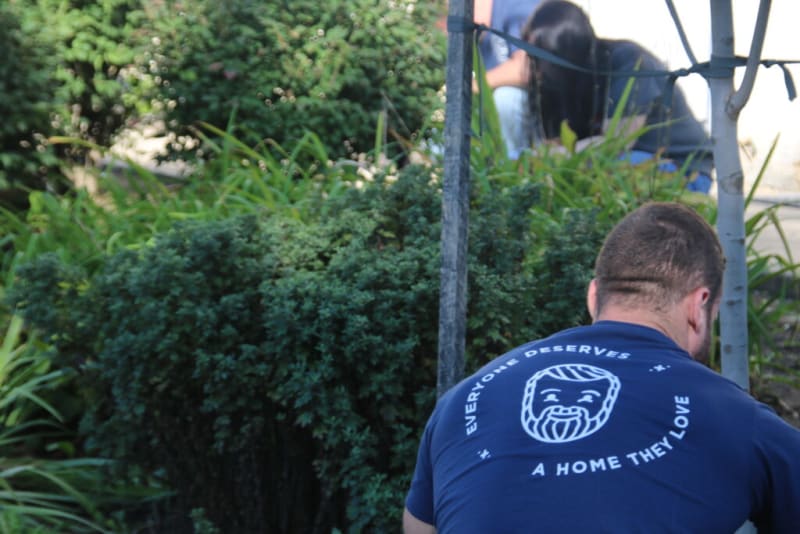 Man wearing a navy blue t-shirt with white text and a bearded face graphic reading "Everyone Deserves A Home They Love," sitting outdoors near greenery.