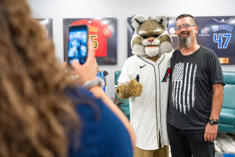 A person posing with the Arizona Diamondbacks mascot wearing a white baseball jersey with the team logo, while another person takes their photo with a smartphone. Background features framed sports jerseys.