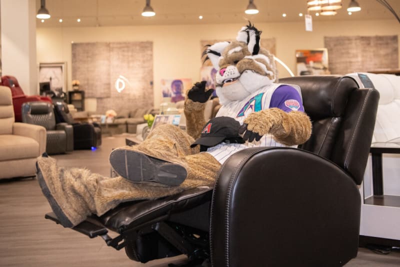A person in a furry mascot costume wearing a white and purple baseball jersey lounges on a black leather recliner chair in a furniture store showroom.