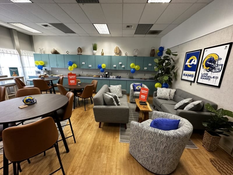 A modern lounge area with gray sofas, patterned chairs, wooden tables, blue and yellow balloons, and Los Angeles Rams posters on the wall.