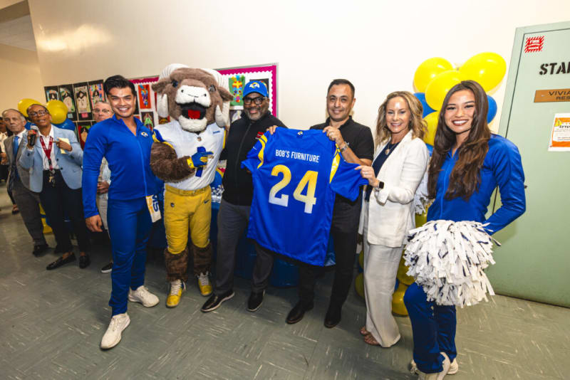 Group of people posing indoors with a mascot holding a blue and yellow sports jersey displaying "BOB'S FURNITURE 24," surrounded by blue and yellow balloons.