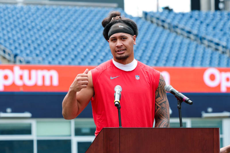 Athlete in a red sleeveless Nike NFL shirt with a tribal tattoo on the right arm, giving a thumbs-up at a stadium podium with microphones.