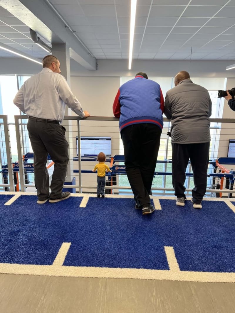 Three men leaning on a railing overlooking a sports training area with blue turf and digital screens, under a grid ceiling with LED lights.