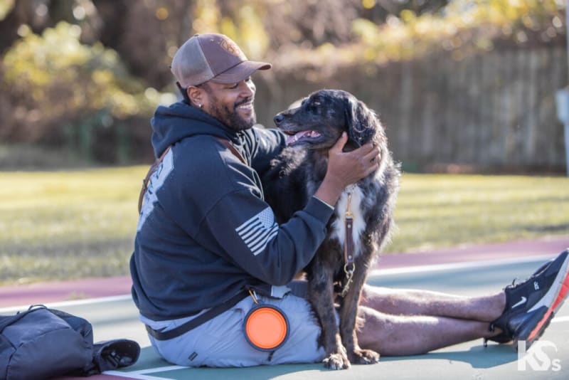 Person sitting on an outdoor court wearing a dark hoodie, gray shorts, and black Nike sneakers, holding a black and brown dog with a leash.