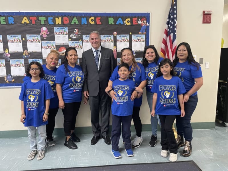 Group of adults and children wearing blue "RAMS HOUSE Inglewood, CA" T-shirts standing indoors near a bulletin board titled "Attendance Race" with colorful letters and an American flag.