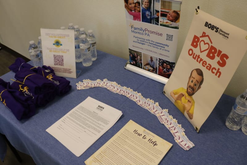 Table display promoting Bob's Discount Furniture outreach and Family Promise Montco PA partnership, featuring purple t-shirts, water bottles, informational flyers, and a sign with a cartoon man making a heart shape with his hands. Text includes "BOB'S Outreach," "Family Promise Montco PA," and details on addressing homelessness and hunger. A QR code and contact info are visible.