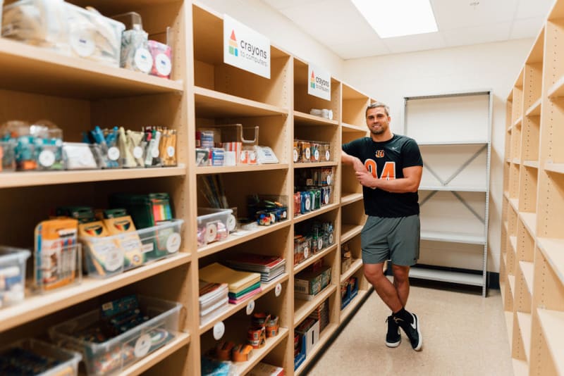 Man standing in an aisle of wooden shelves stocked with art supplies like crayons, markers, notebooks, and craft materials labeled "crayons to computers."