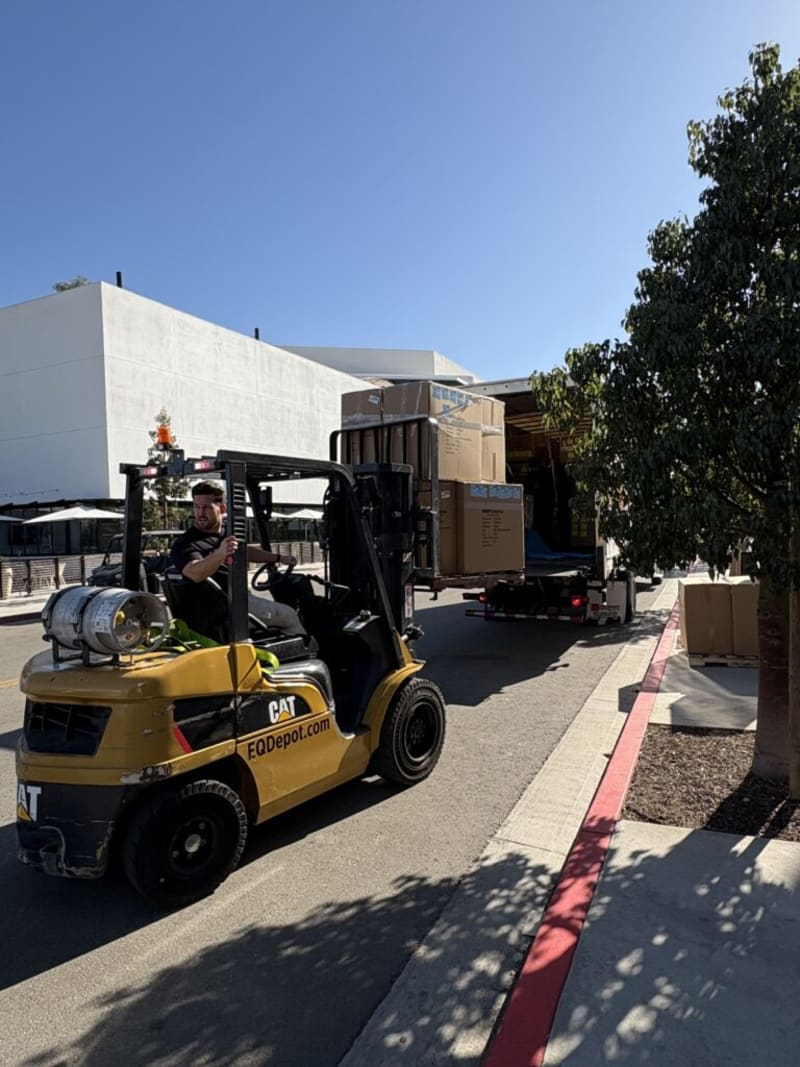 Yellow CAT forklift with EQDepot.com branding lifting stacked cardboard boxes from a delivery truck on a sunny street near a sidewalk and tree.