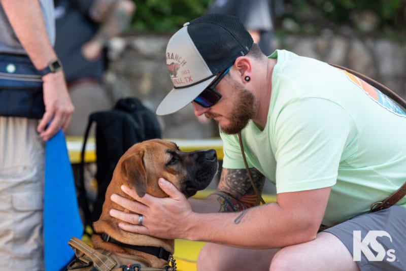 Man wearing a light green t-shirt, gray shorts, and a trucker hat gently holding a brown service dog with a harness, outdoors.