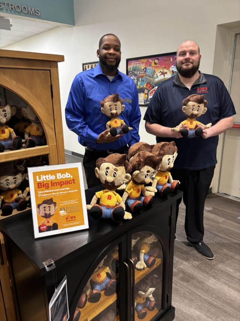 Two men standing in a store holding plush toys of a cartoon character named Little Bob, wearing yellow shirts and blue pants. A display cabinet holds multiple identical Little Bob plush toys. A framed sign reads: "Little Bob, Big Impact. Take Little Bob home and help Children's Miracle Network Hospitals. $11.99." The setting promotes Little Bob plush toys supporting charity through Boss's Furniture.