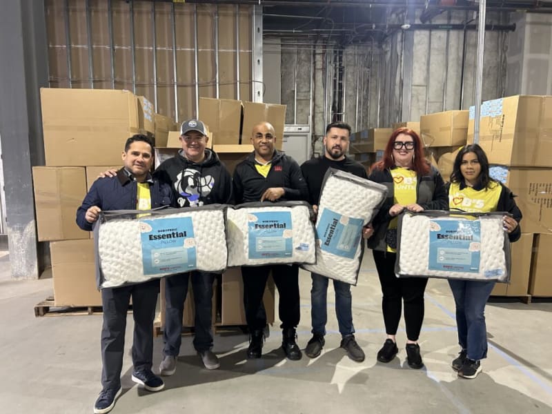 Six people standing in a warehouse holding packaged BOBSPEDIC Essential Pillows with white quilted texture and blue labels, surrounded by stacked cardboard boxes.
